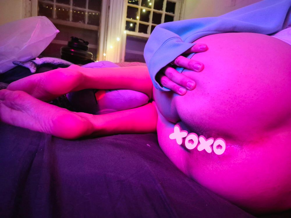 A bright pink peach lying on a bed, spreading cheeks with one hand. Their knees are curled up so their fee5 and calves are exposed