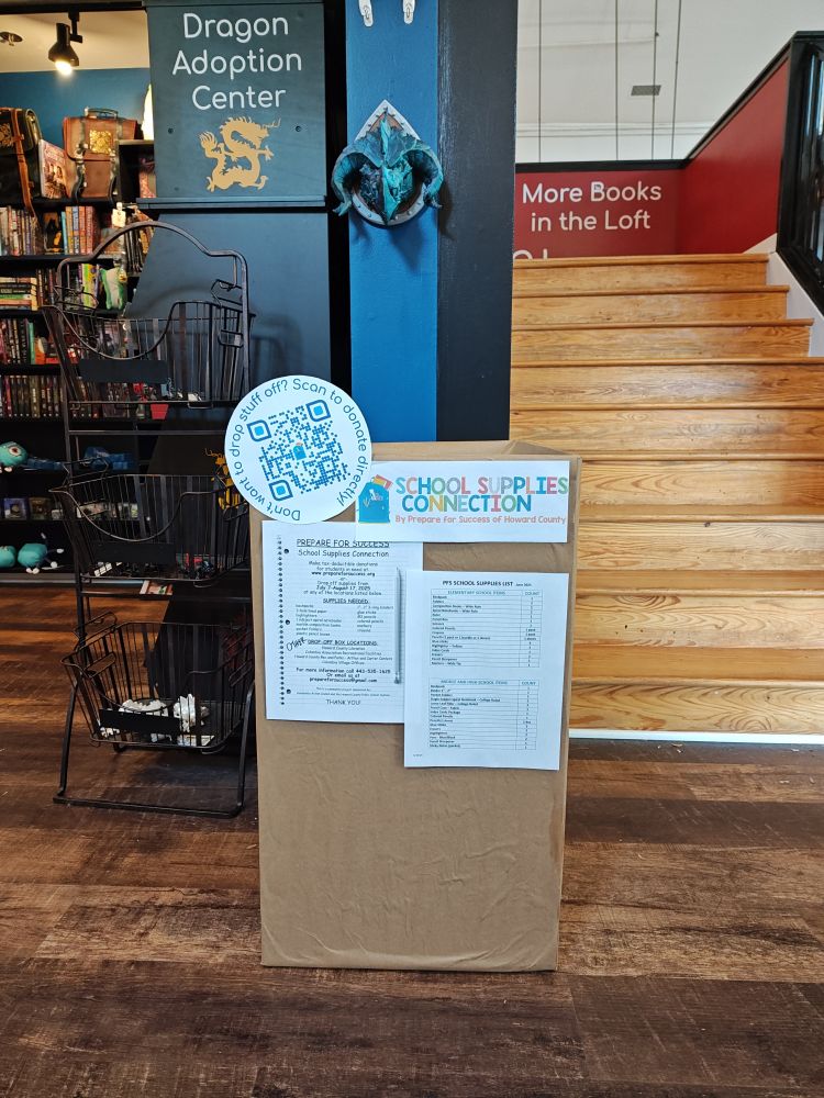 Image of a large box marked with the logo of Prepared for Success's School Supply Connection program. There are school supply lists affixed to the box. The photo is framed to show where the box is located in the store: next to the Dragon Adoption Center at the foot of the stairs.