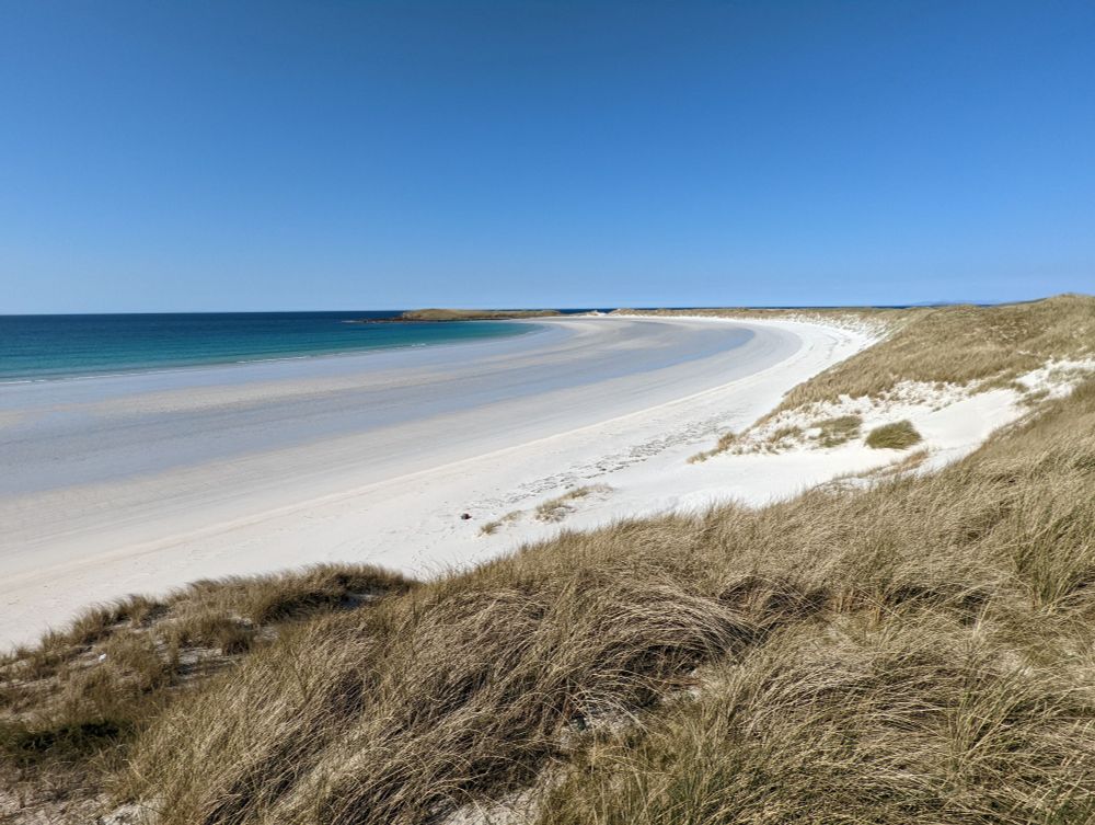 A deserted sweeping bay with pale sandy beach backed by high sand dunes covered in Marram grass. The sea is dark turquoise to Prussian blue in the sunlight.