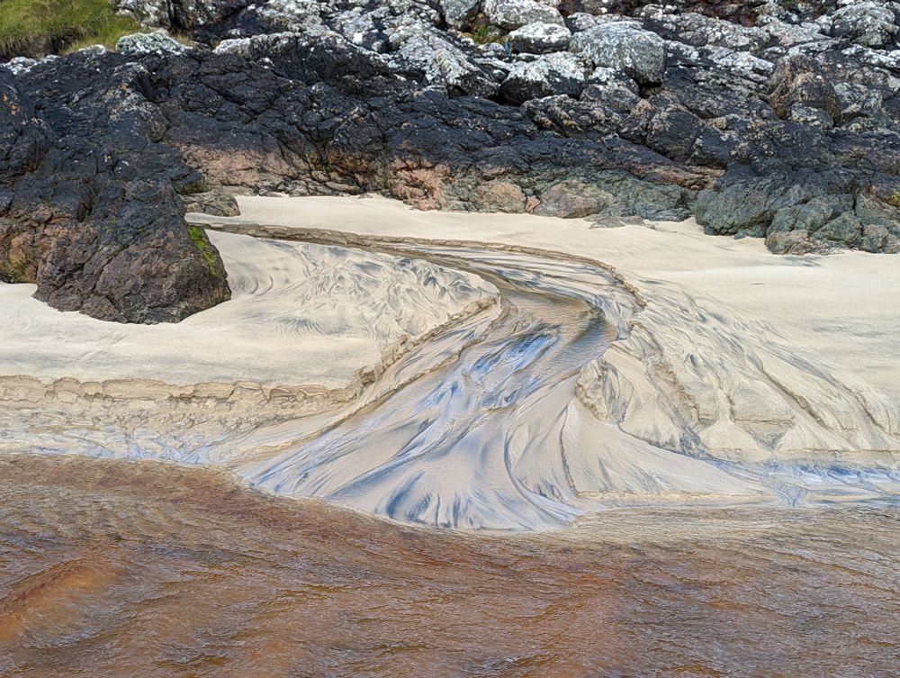 Small tributary stream building a delta into larger brown, (peat stained) stream.  The sedimentary features observed in the yellow sand are emphasised by black heavy mineral concentrations.
