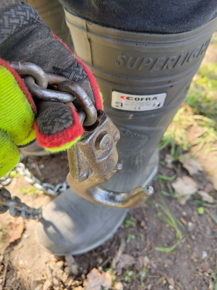 hook of a chain hoist held in front of a black Cofra rubber boot by a hand wearing a red and yellow work glove,