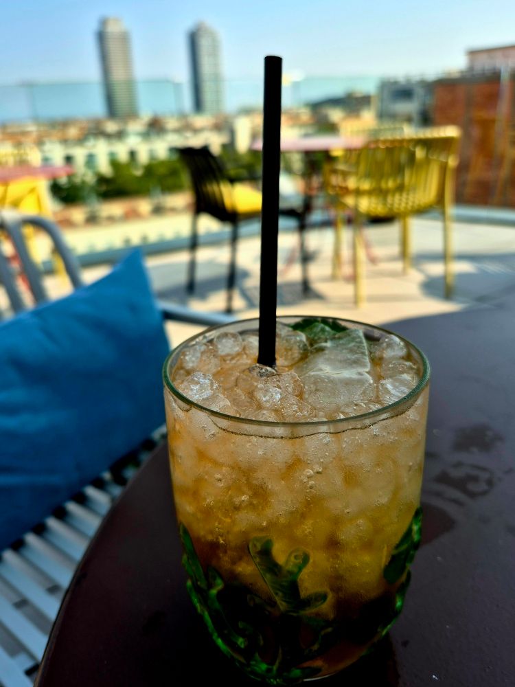 Glas Mojito gefüllt mit Eiswürfeln, Minzblättern und einem schwarzen Strohhalm. Im Hintergrund ist eine Dachterrasse mit Stühlen und Tischen zu sehen, dahinter Stadtlandschaft