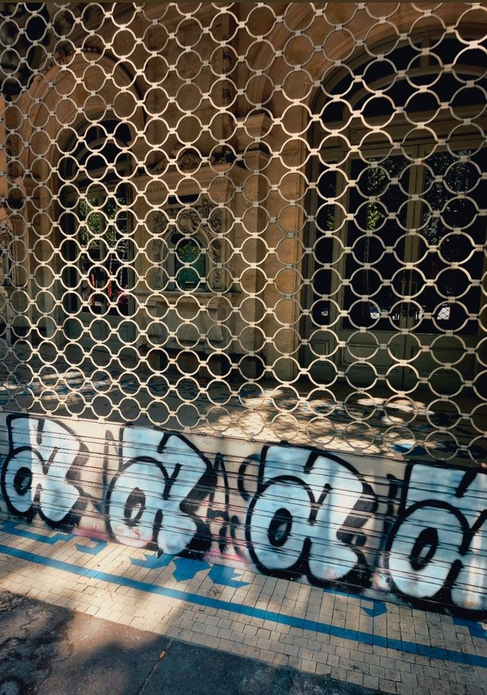 ​Ein geschlossenes Ladenlokal mit einem Metallgitter davor. Unten am Gitter ist ein großes Graffiti mit weiß umrandeten schwarzen "a" Kleinbuchstaben zu sehen.