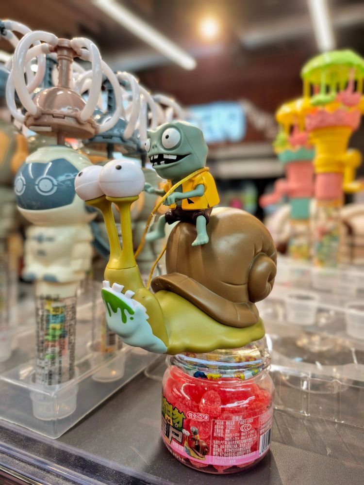 ​Die Szene zeigt eine Spielfigur aus dem Videospiel "Plants vs. Zombies". Es ist ein Zombie mit gelbem Hemd und blauen Shorts, der auf einer großen Schnecke reitet. Die Schnecke hat Augen am Ende von Stielen, eine helle, grün-gelbe Schale und tropfenden Schleim am Mund. Die ganze Figur steht auf einem durchsichtigen Glas mit Süßigkeiten, das auf einem Regal in einem Geschäft steht. Im Hintergrund sind weitere ähnliche Figuren zu erkennen.