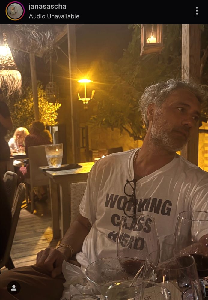 An Instagram photo by user Janasascha of Taika at an outdoor restaurant at night. He's wearing a white tee with the words "Working Class Hero" on it and looking out of frame at someone. A long bit of his neck is exposed and his glasses are hanging from his shirt collar, where you can see a peek of gold necklace. There are lots of empty wine glasses on the table in front of him. 