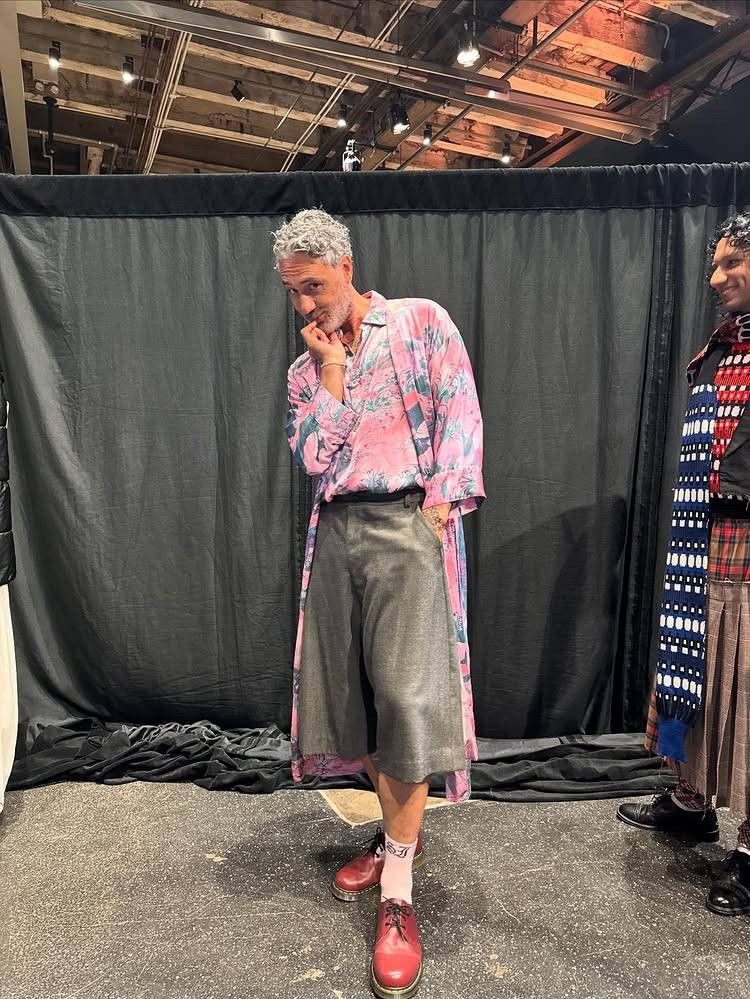 Taika Waititi from 2025 Paris fashion week behind the scenes. He's in a pink floral shirt with a matching pink floral silk robe. His trousers are gray and just below knee-length and he has red shoes and crew socks on. He's standing in front of a black fabric privacy curtain in what looks like a giant warehouse. 