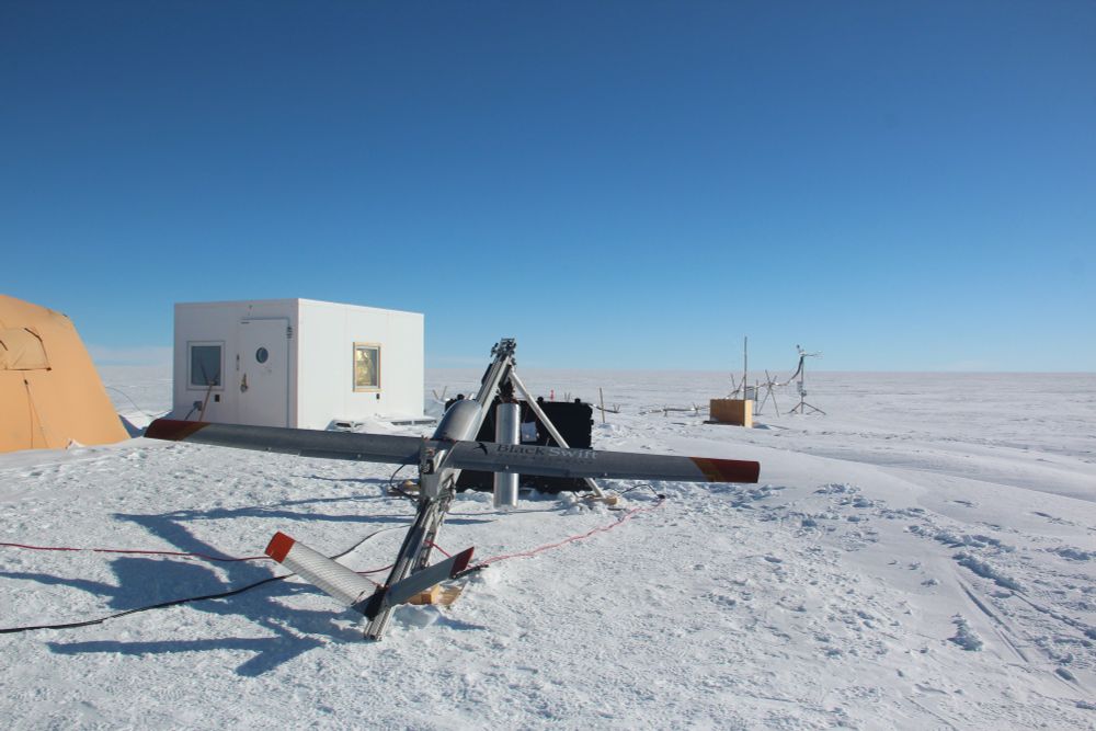 Picture of a measurement site on the Greenland Ice Sheet. In the foreground is a fixed‐wing uncrewed aerial system (UAS), looking like a small, light plane with a wing span of 3 m. The UAS is prepared for take-off on a launching system with a high pressure flask that is filled with air through a connected compressor. In the background are white, heated measurement cabin and a turbulence measurement system (eddy covariance) that is connected with a thick bundle of cables to the cabin. 