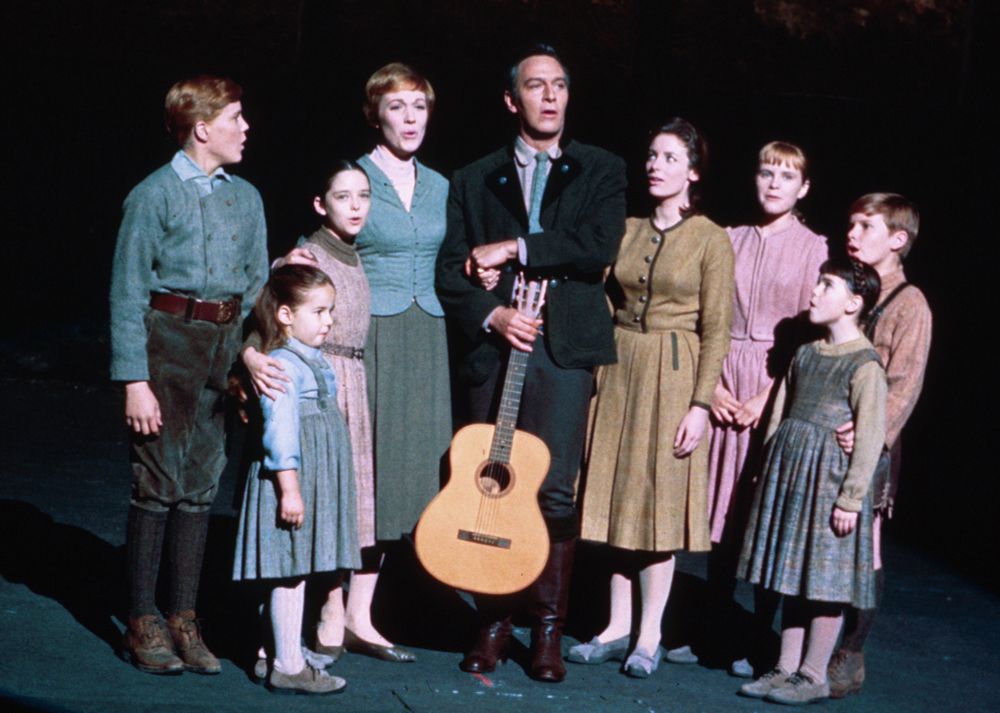 A family stands on a stage. The adult male in the center holds a guitar. The rest of the family stands closely to him and each other as they sing. The von Trapp Family. 

Image from "The Sound of Music" (1965). This image is from the scene when they are made to perform for a large group of Nazis. Shortly after, the family escapes from the Nazis and leaves the country.