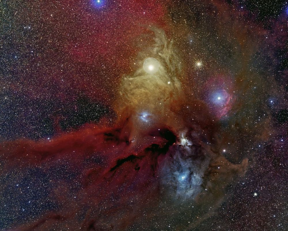 "Colorful wide-field image of the Rho Ophiuchi cloud complex, featuring vivid hues of blue, orange, red, and brown. Wispy reflection nebulae intertwine with dark dust lanes and glowing hydrogen clouds, creating a rich, high-contrast tapestry of interstellar gas and dust. Bright stars Antares and Rho Ophiuchi are prominent amid the surrounding cosmic material."