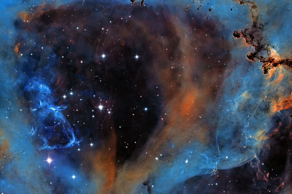 A high-resolution narrowband image of the core of the Rosette Nebula (NGC 2244), featuring vivid hues of blue and orange. Central dark dust lanes are surrounded by intricate structures of ionized gas glowing from nearby young stars. The image showcases fine detail and high contrast between light and shadow in space.

