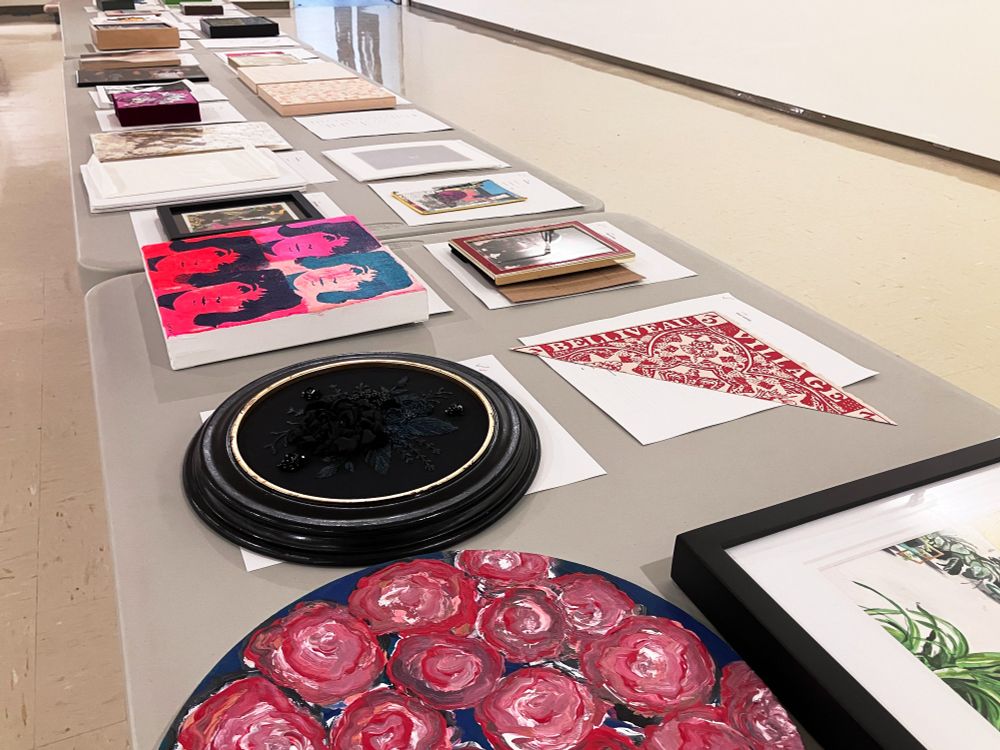 Dozens of artworks are laid out on a long line of grey tables. 