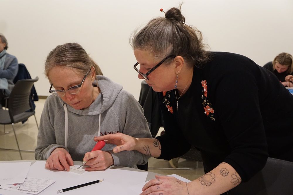 Tara leans in to help a workshop participant as they carve an eraser stamp.