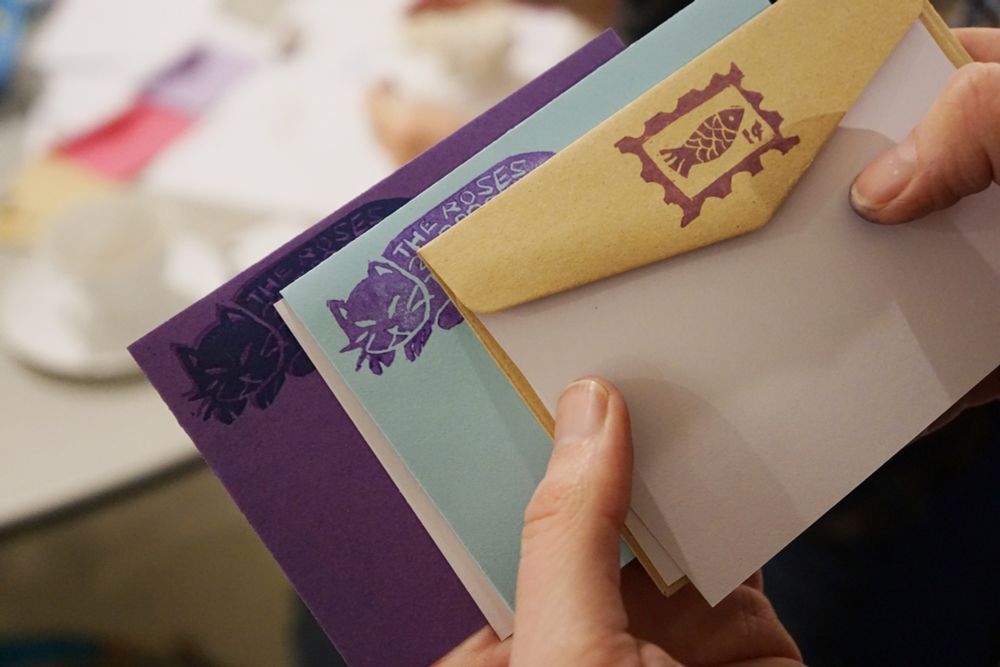 Two hands hold a selection of envelopes. Two contain a cat stamp with return address information and the one on top has a purple stamp of a postage stamp with a fish motif.