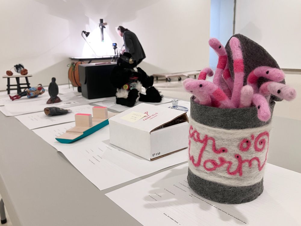 Artworks, including a felted can with worms are displayed on a grey table. In the background a person works at a copy stand.
