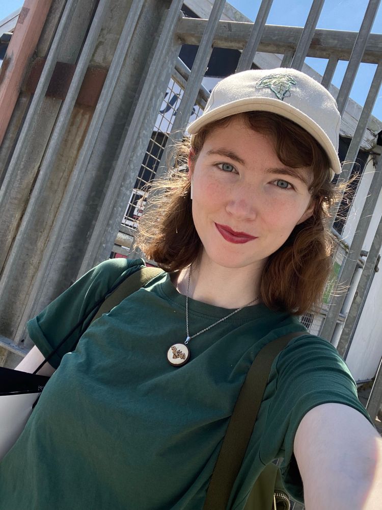 Selfie of SB wearing a green T-shirt and a beige hat with a hand embroidered outline of a leaf of Lórien from Lord of the Rings, and an embroidered bat necklace. Her hair is down and she is wearing a deep pink lipstick 