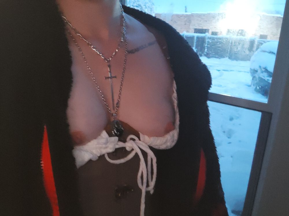 Landscape shot of a person's upper torso with small breasts exposed. White top is pulled down for hard nipples to be visible above a brown corset. Person wears a black fluffy jacket and silver necklaces and stands in front of a half open screen door with heavy snow outside. 