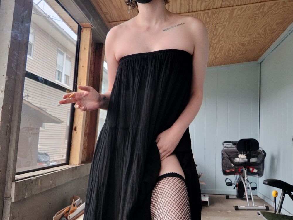 Ryder wearing a long black skirt as a dress and pulling up the hem to show fishnet thigh highs. He holds a cigarette in one hand
