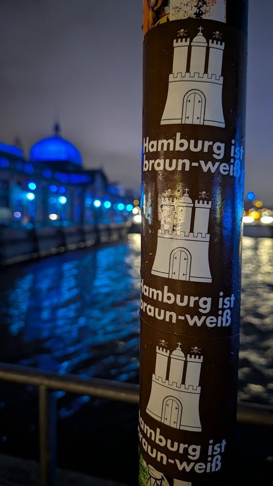 Ein Laternenpfahl am Hamburger Hafen, beklebt mit mehreren Stickern in Braun mit dem weißen Hamburger Stadtwappen und dem Schriftzug „Hamburg ist braun-weiß“ – ein Hinweis auf die Vereinsfarben des FC St. Pauli. Im Hintergrund leuchten Gebäude und Wasserflächen in intensivem Blau bei Nacht.