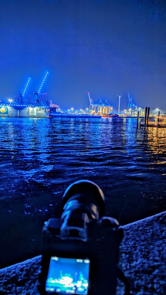 Kamera auf einem Ministativ unscharf im Vordergrund, dahinter blau beleuchtete Hafenkräne und Brücken anlässlich des "Blieb Harbor"
