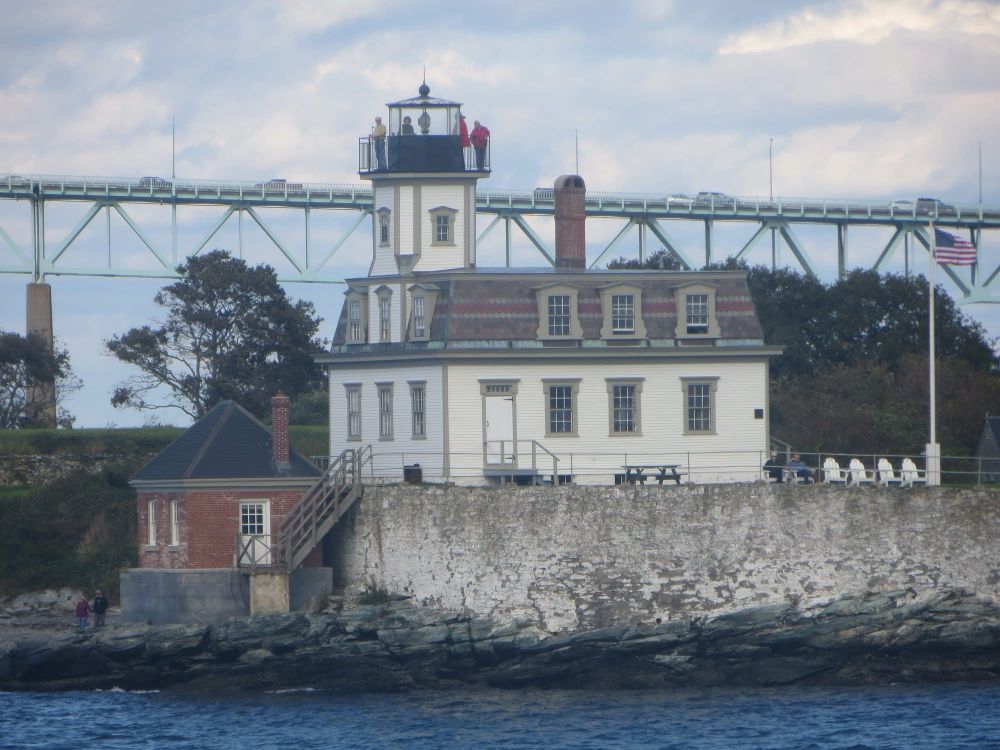 A two-story wooden house with gabled windows and a mansard roof stands on a large masonry foundation. A short square tower topped with an iron lantern and a surrounding walkway rises from the front section of the roof. People are standing inside the lantern room and on the walkway. A US flag flies from a pole to the left of the lighthouse. A high bridge with cars driving back and forth is in the background, and the calm, deep-blue water of the harbor is in the foreground. The sky is mostly cloudy, with patches of blue.