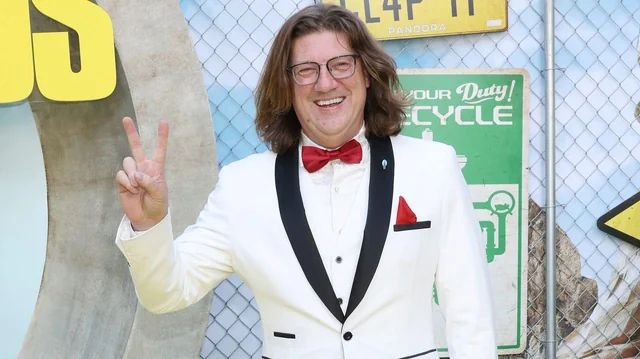 this is a picture of randy pitchford holding up a 'peace' sign with his fingers at a press event. he is grinning, he looks like a tool, his suit is awful