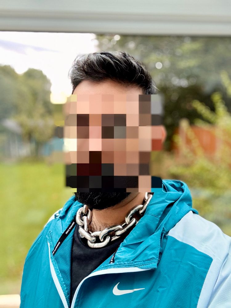 An identity obscured male wearing a thick marine chain and a Nike running jacket. The background is outdoors but blurred. 