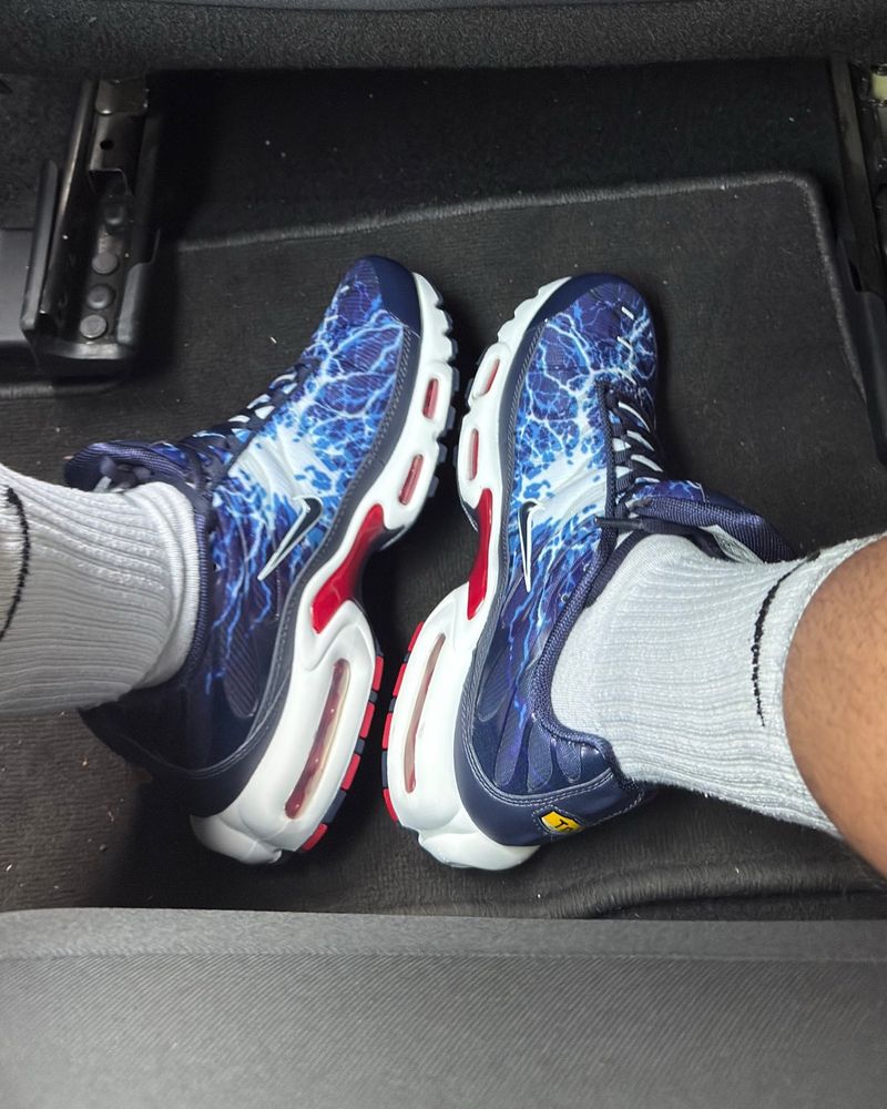 A pair of Nike TN Eclair Lightning pattern in blue white and red. Nike socks. Hint of a thick calf. In the back seat well of a car. 