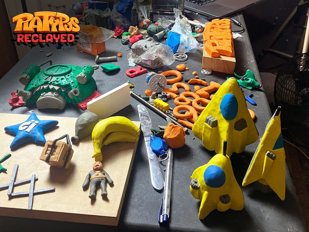 The image depicts a messy desk of clay sculptures made for Platypus Reclayed. Visible are models of numbers, player and enemy ships, bananas, star power-up, crate, balloon, enemy pilot, game logo, etc. Platypus Reclayed (2025) is an upcoming remake of Platypus (2002). Both are claymation shoot 'em up games.