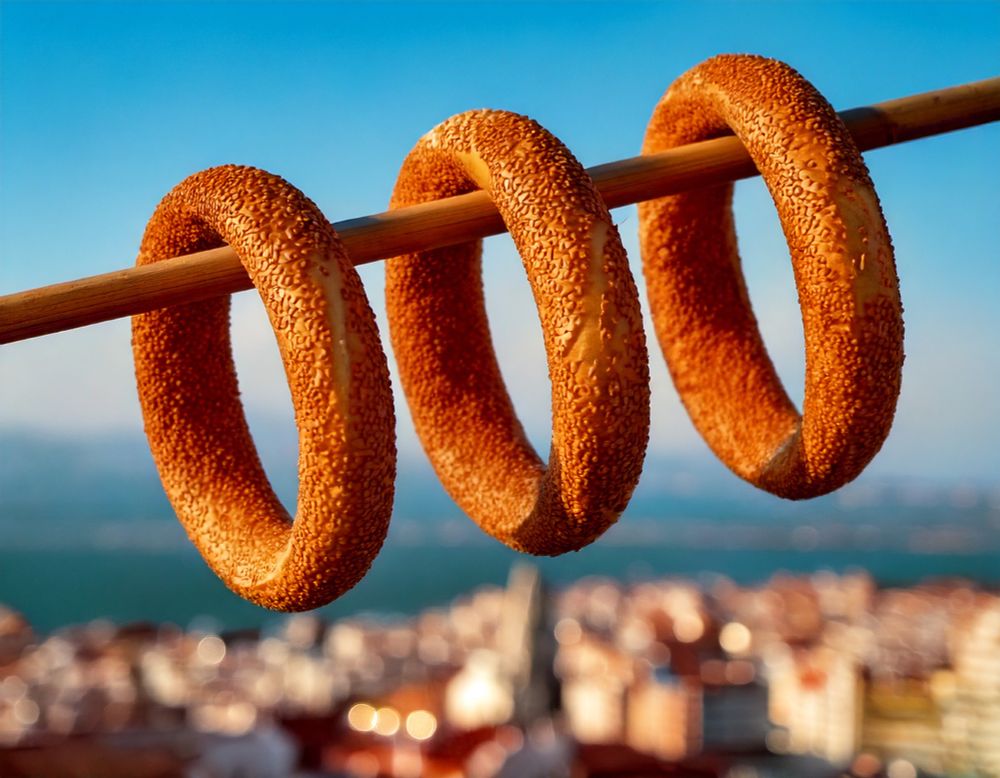 Three golden, sesame-coated koulouri (a traditional Greek bread ring) hanging on a bamboo rod. The image captures the warm glow of a sunset or sunrise with a blurred city and waterfront in the background, highlighting the vibrant golden tones of the simit.