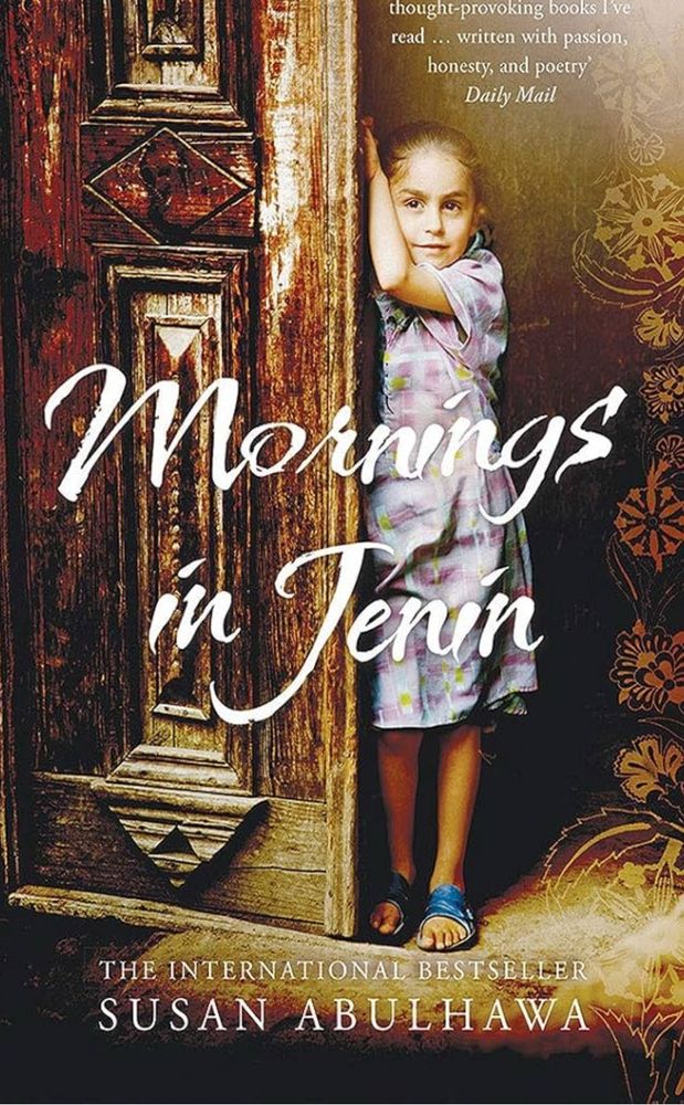 Cover image of the Book Mornings in Jenin shows a young girl standing against a large, worn wooden door with intricate moulding patterns. She wears a lilac dress that goes to her knees
And blue sandals. Her brown hair is pulled back and one stem is up holding the edge of the door. 