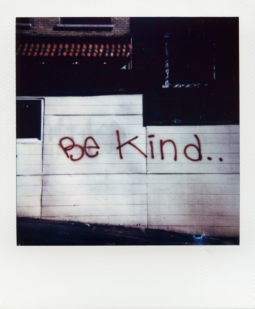 Polaroid picture. The whitewashed wooden wall of a parklet has been tagged in red with the message "Be Kind.." I don't know why there are two dots instead of a period or an ellipses.