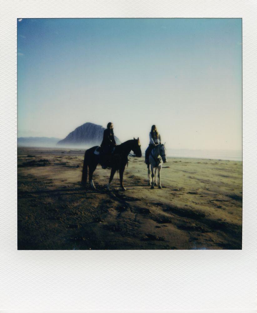 Polaroid picture of two teenage girls on horseback on the beach with Morro Rock in the distance. The late day light is low and golden, casting long shadows.