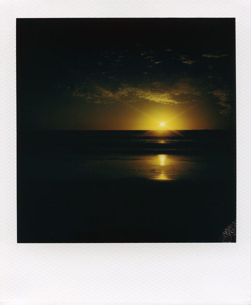 A polaroid picture of the sun setting over the Pacific ocean. The golden sun sends out rays of light and reflects on the surf and sand below and illuminates the clouds above.