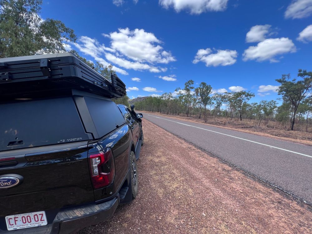 A Black Ford Next Gen Ranger is on the side of a remote highway. It has a rooftop tent affixed. The sky is blue with a scattering white clouds. The earth, a reddy brown. Green-leaved bush trees pepper the highway. Location: Remote Northern Territory, Australia.
