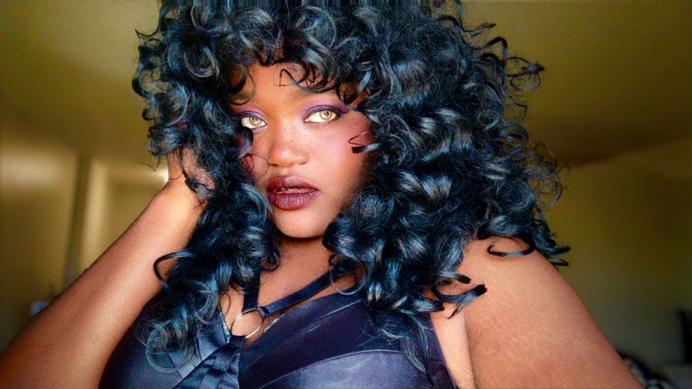 A picture of Bree in black curly hair with makeup on. They have a “fierce” expression on her face.
