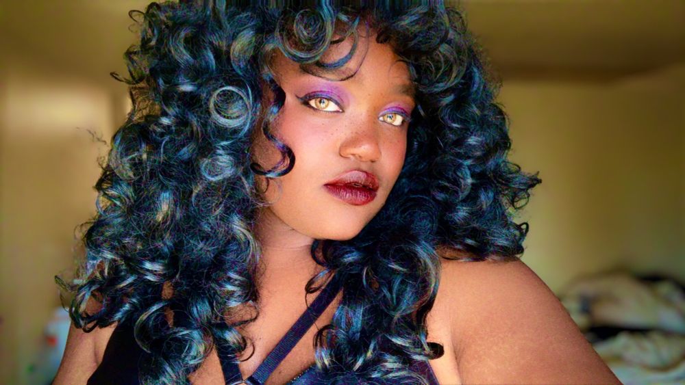 A picture of Bree in black curly hair with makeup on and a neutral expression. 
