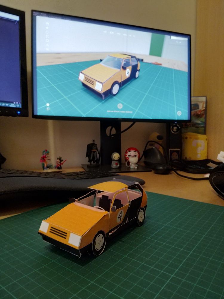 A real black & yellow car made out of cardstock sits on top of a green cutting mat on a desktop. Behind it there's a PC monitor showing the exact same car inside the videogame. An ergonomic keyboard and some figurines are also visible.