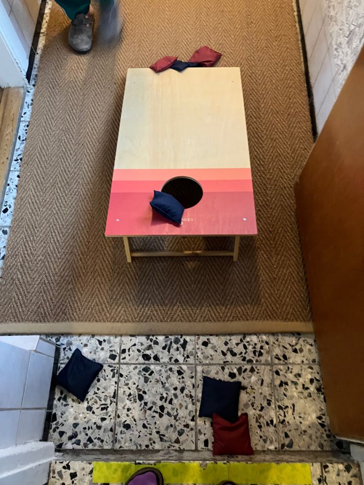 Ein Cornholebrett (das ist ein Brett, das leicht schräg geneigt auf den Boden gelegt wird und bei dem Versucht werden muss ein Loch mit einem Säckchen zu treffen, welches ursprünglich mit Maiskörnern gefüllt war) 
Das Brett steht auf einem Steinboden auf dem ein Läufer aus Bast liegt. Darauf und daneben liegen blaue und rote Säckchen