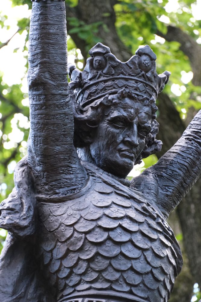 A zoomed-in photograph of the Polish King Jagiello statue in Central Park.  The statue is wearing a crown and a scaled breastplate, with his arms raised up high above him.  His head is tilted down as he stares ahead intensely.