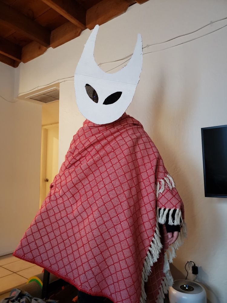 A person wearing a cardboard mask mimicking Hornet from Silksong, they are also wearing a red blanket wrapped around their shoulders as her cloak. 