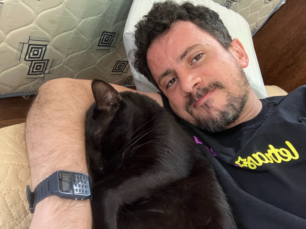 Homem deitado na cama com gato aconchegado no braço, de olhos fechados.