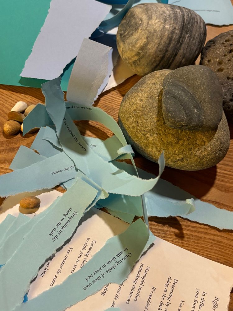 Scraps of blue, purple and white torn paper with fragments of poems on them curling on a wooden surface next to four grey stones. 