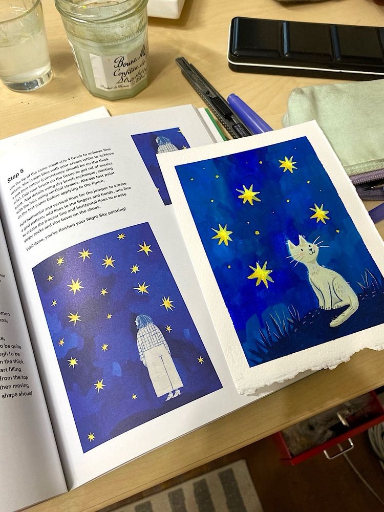 Gouache painting of a white cat against a blue sky with yellow stars. To the left is a book with a painting of a woman in white against blue sky and yellow stars. The cat painting is based on a tutorial in the book.