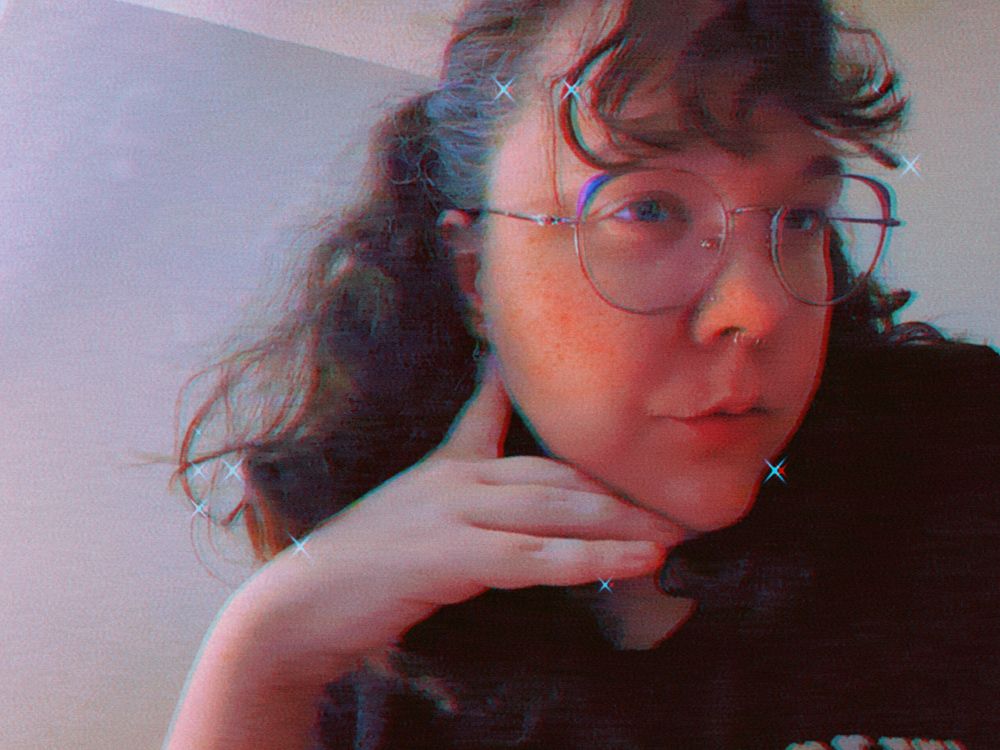selfie from the chest up; i'm a white woman wearing large round glasses with purple at the upper outer corners. my hair is curly and brown and the upper half has been pulled up into two little pigtails. i'm resting my chin on my hand and looking at myself on the phone screen instead of into the lens. the image is filtered; chromatic aberration and sparkles have been applied.