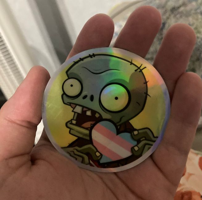 A hand holding a sticker of a PvZ 2 zombie holding a heart with the trans flag design inside it.

Image taken by @HATCHPANEL on X/Twitter.
