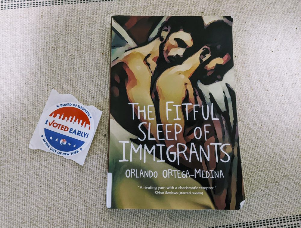 Photo of the book "The Fitful Sleep of Immigrants" whose cover is drawn art of two men holding each other; plus an "I Voted Early" sticker.