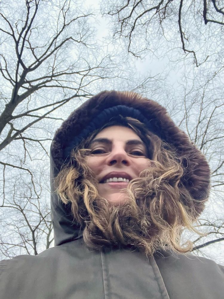 A selfie taken from a low angle showing a person wearing a thick hooded coat lined with fur, smiling against a backdrop of bare winter trees and an overcast sky. The person looks warmly wrapped up for a cold day.