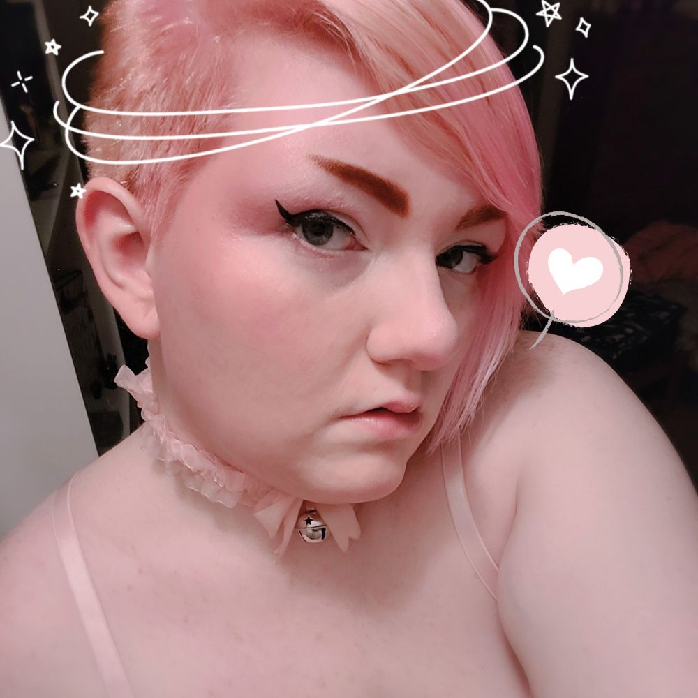 A selfie I took where my head is slightly tilted to the side and downward. I'm wearing a lacy collar with a bell. There are stickers of a heart inside a speech bubble, and a halo around my head. 