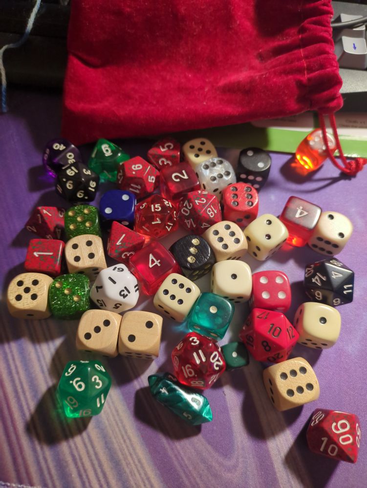 Ein Haufen verschiedener Würfel in unterschiedlichen Farben und Größen mit einem roten Säckchen im Hintergrund 