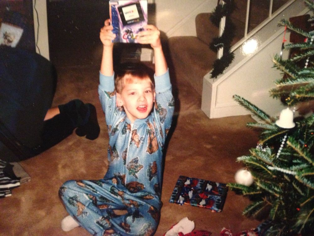 Caleb as an elementary aged kid on Christmas in the late 90s/early 00s, displaying a purple translucent GameBoy Color box he received as a present. He is wearing blue fleece pajamas with brown bears on them and he is sitting next to a Christmas tree